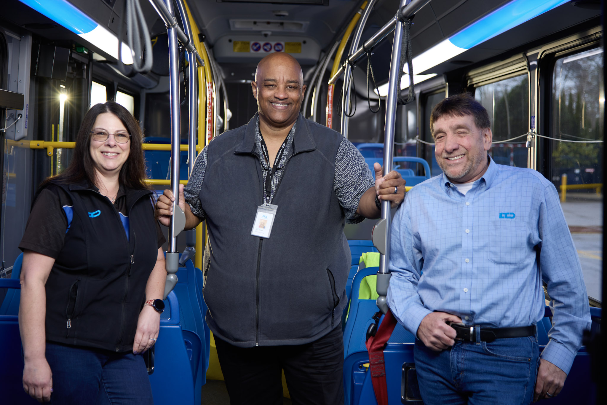 Three people stand and smile inside a city bus, holding onto poles. The bus seats and interior are visible in the background.
