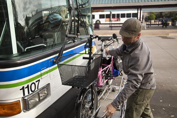 A man loads a bicycle with a front basket onto the bike rack of a city bus at an outdoor transit station.