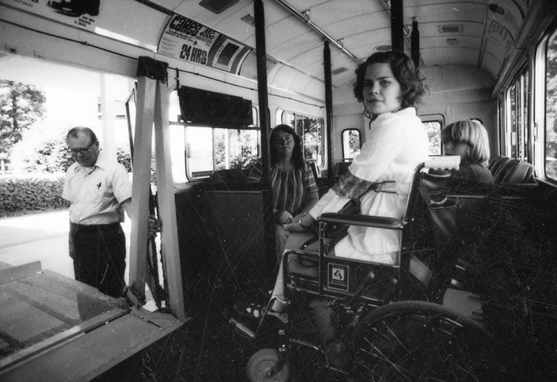 A woman in a wheelchair is positioned on a bus lift while two women sit nearby and a man stands outside the bus door.