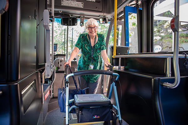 An older woman stands inside a bus, smiling, with both hands on a walker. Daylight comes through the windows behind her.