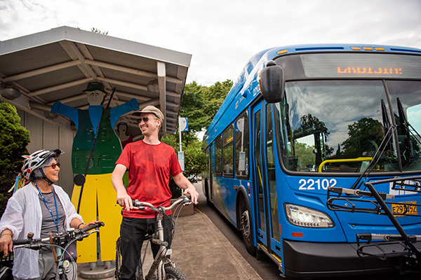 Two people with bicycles stand near a blue bus at a bus stop, smiling at each other. There is a large cutout figure behind them and trees in the background.
