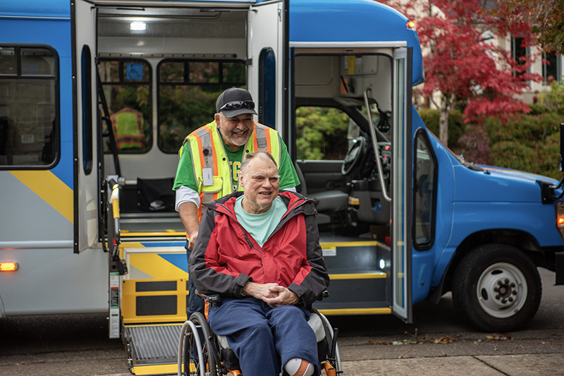 A man in a wheelchair is being assisted by a transit worker onto a blue accessible van using a wheelchair lift, with trees visible in the background.