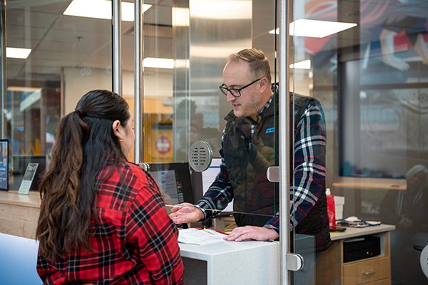 A man speaks with a woman at a service counter inside an office, separated by a glass barrier.