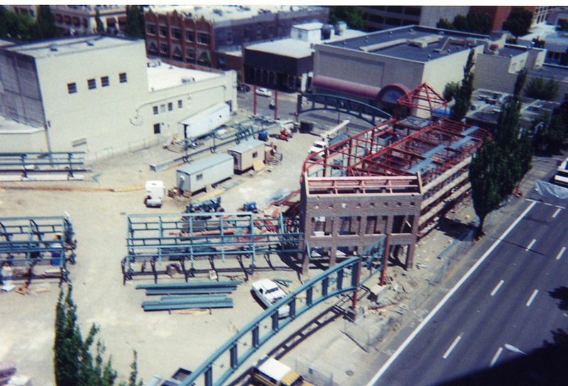 Aerial view of a construction site with steel framework, a partially built brick structure, equipment, and nearby city streets and buildings.