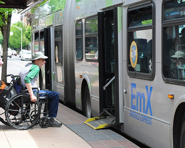 A man in a wheelchair waits to board a city bus using a deployed accessibility ramp at a bus stop.