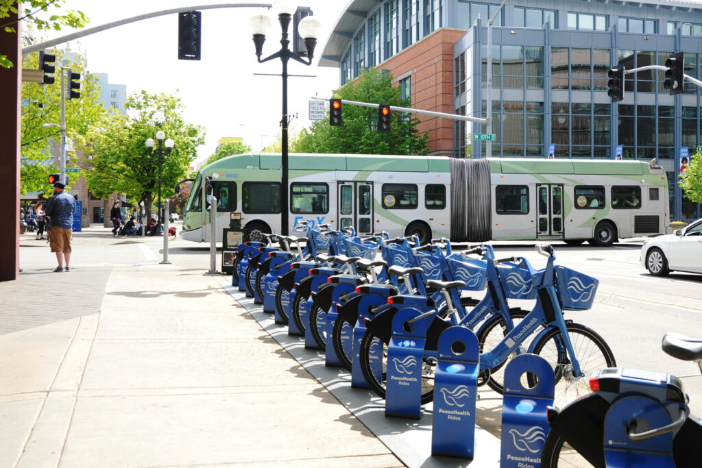 A row of blue rental bikes is lined up at a dock on a city sidewalk; a green city bus passes by in the background.