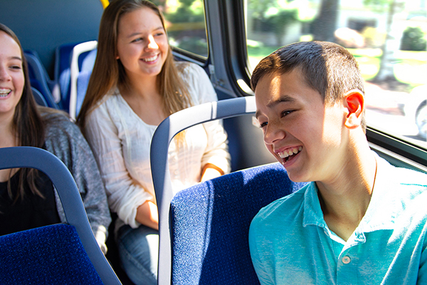 Three teenagers sit together on a bus, smiling and talking while sunlight streams through the window.