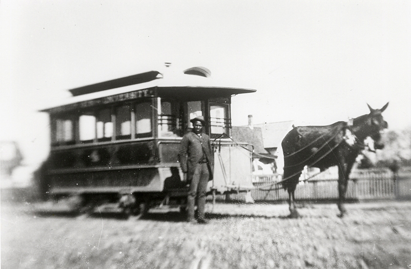 A man stands next to a mule-drawn streetcar on a dirt road, with buildings and a fence in the background.