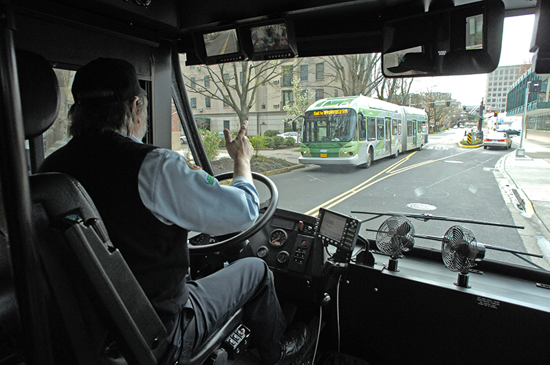 A bus driver gestures with his hand while driving, as another green city bus approaches in the opposite lane on a city street.