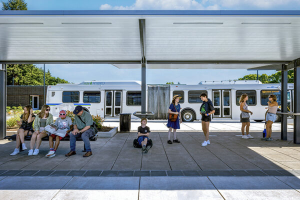 People wait at an outdoor bus stop under a shelter, with a white hybrid bus parked in the background on a sunny day.