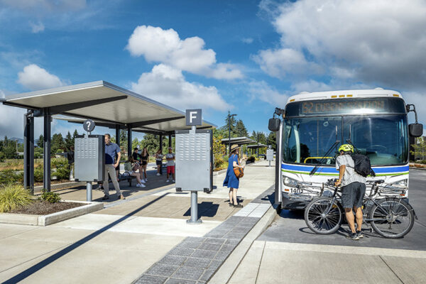 People wait at a covered bus stop while a cyclist loads his bicycle onto the front rack of a bus numbered 62 on a sunny day.