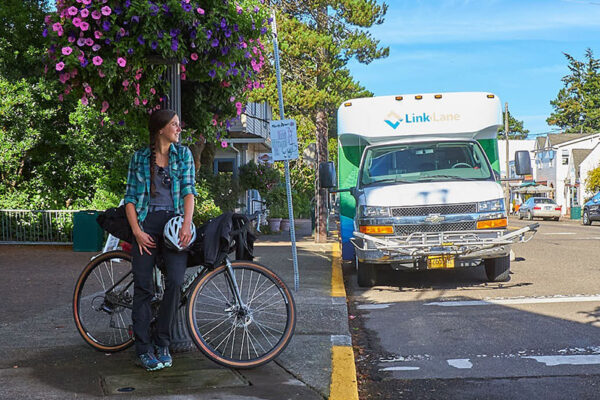 A person sits on a bicycle near a sidewalk, holding a helmet, with a Link Lane shuttle bus parked on the street in the background.