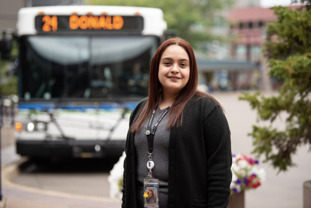 A woman wearing a black cardigan stands outside in front of a city bus displaying the route “21 DONALD.”.