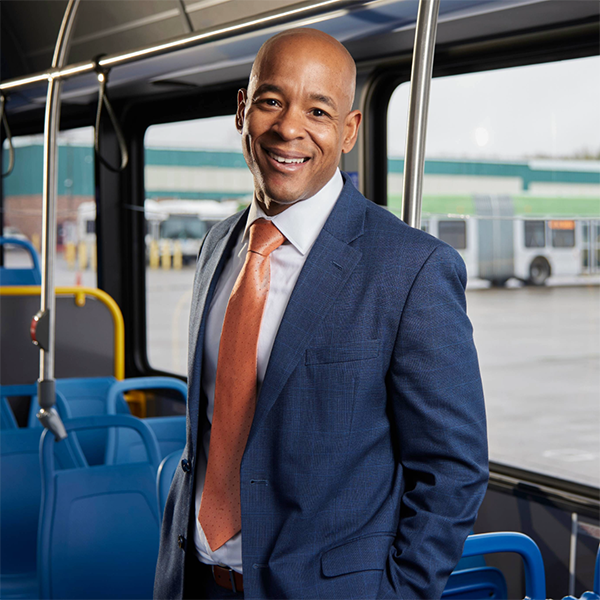 Jameson T Auten, CEO of Lane Transit. A man in a blue suit and orange tie stands smiling inside a city bus, with bus seats and a window showing another bus and a building outside.