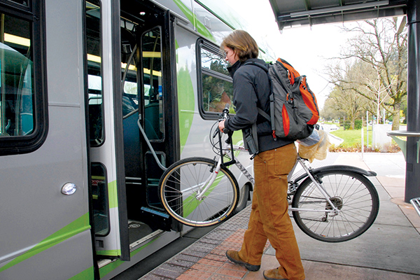 Person boarding a bus while carrying a bicycle, wearing a backpack and brown pants, at an outdoor transit stop.