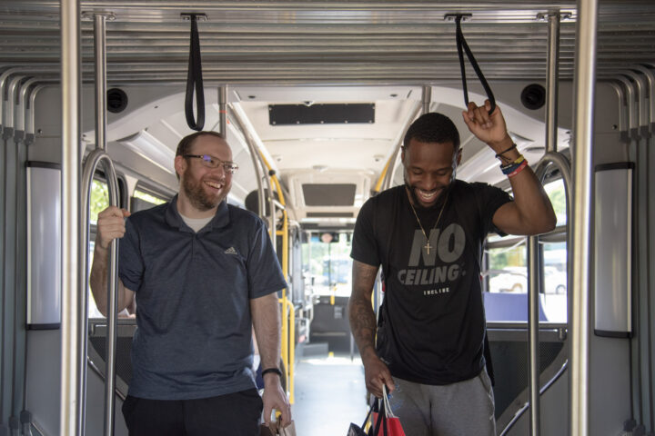 Two men stand inside a city bus, holding onto hand straps. They are smiling and appear to be engaged in conversation.