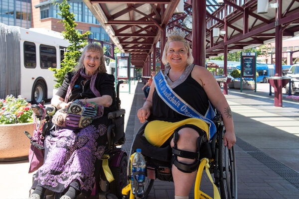 Two women in wheelchairs, one wearing a sash and crown, sit together outdoors at a transit station under a red canopy.