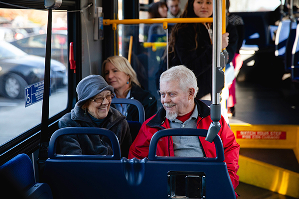 Two older adults sit and smile at each other on a city bus, with other passengers visible in the background.