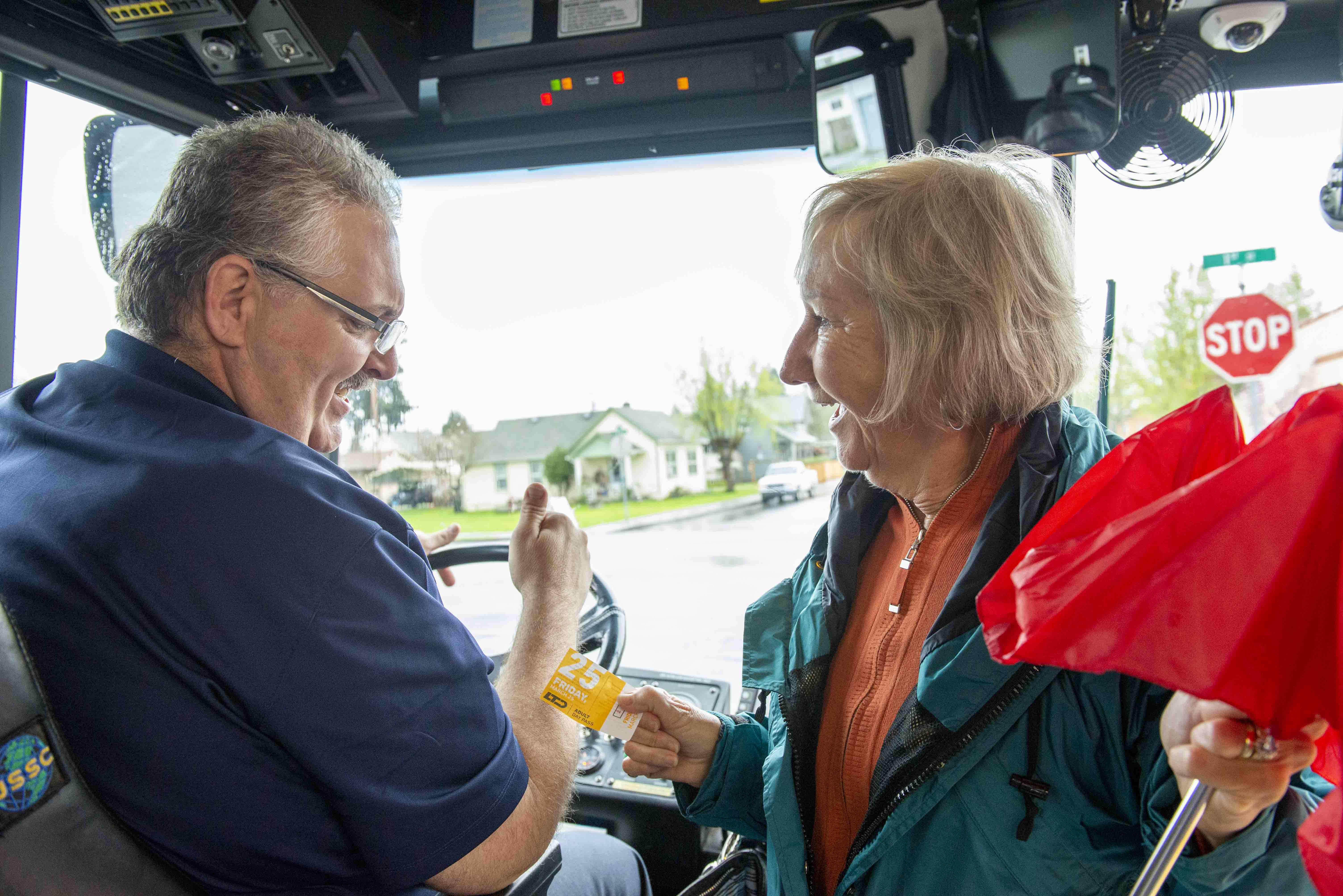 An older woman hands a yellow bus ticket to a male bus driver while holding a red umbrella, both smiling inside a bus.