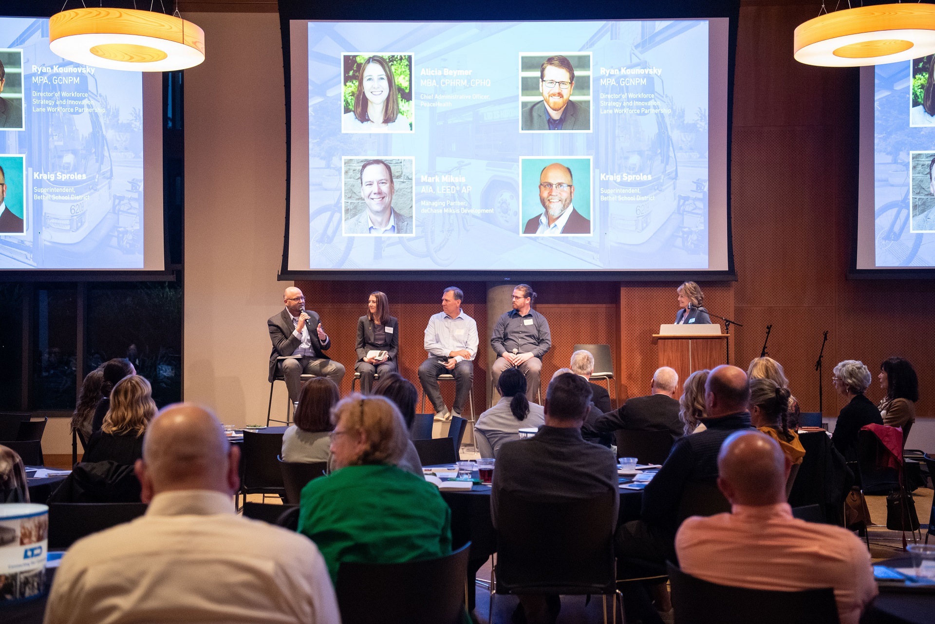 A panel of five speakers sits on stage in front of an audience, with presentation slides featuring their portraits and names displayed on large screens behind them.