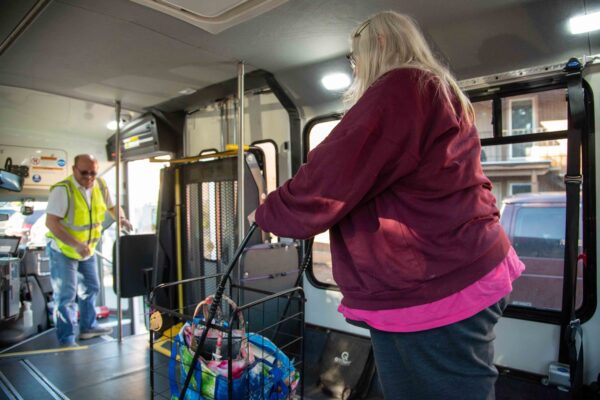 A woman with a shopping cart boards a bus while a man in a yellow vest stands near the entrance.